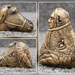 May include: A brass-colored metal object featuring a detailed relief sculpture of a horse head on one side and a human figure wearing a hooded garment on the other. The piece has a hinged construction, showing a textured finish and metallic sheen.