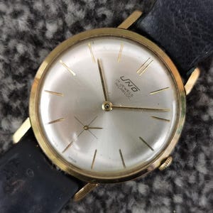 Op de afbeelding: Een vintage goudkleurig polshorloge met een zilveren wijzerplaat en een zwarte leren band. De wijzerplaat heeft goudkleurige uurmarkeringen en wijzers. Het horloge is gemarkeerd met "UNO" en "17 JEWELS INCABLOC".