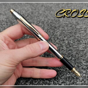 May include: A silver and gold Cross fountain pen with a black grip and gold nib. The pen is held in a hand and the word "CROSS" is displayed in gold lettering.