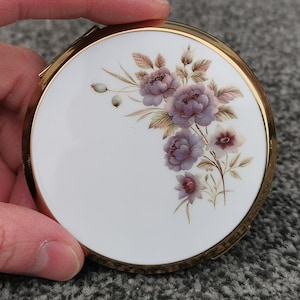 Puede incluir: Un estuche compacto redondo y vintage con un marco de metal dorado. La tapa blanca presenta un diseño floral con flores moradas y rosas y hojas verdes. El estuche compacto está sostenido por una mano.