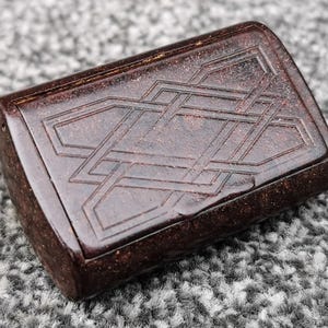 Puede incluir: Una caja rectangular de color marrón oscuro con un diseño geométrico entrelazado tallado en la parte superior. La caja tiene una forma ligeramente redondeada y una superficie lisa y pulida. El intrincado patrón añade un toque de elegancia al pequeño contenedor.