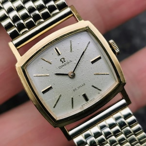 May include: A vintage Omega De Ville wristwatch with a gold-toned square case and a silver-colored dial. The watch features gold-toned hands and markers, and a mesh-style gold-toned band. The watch face displays the Omega logo and the words "De Ville".