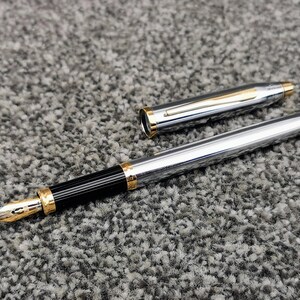 May include: A silver fountain pen with gold accents and a black grip. The pen is uncapped, revealing the gold nib. The cap is lying next to the pen. The pen is on a gray, textured surface.