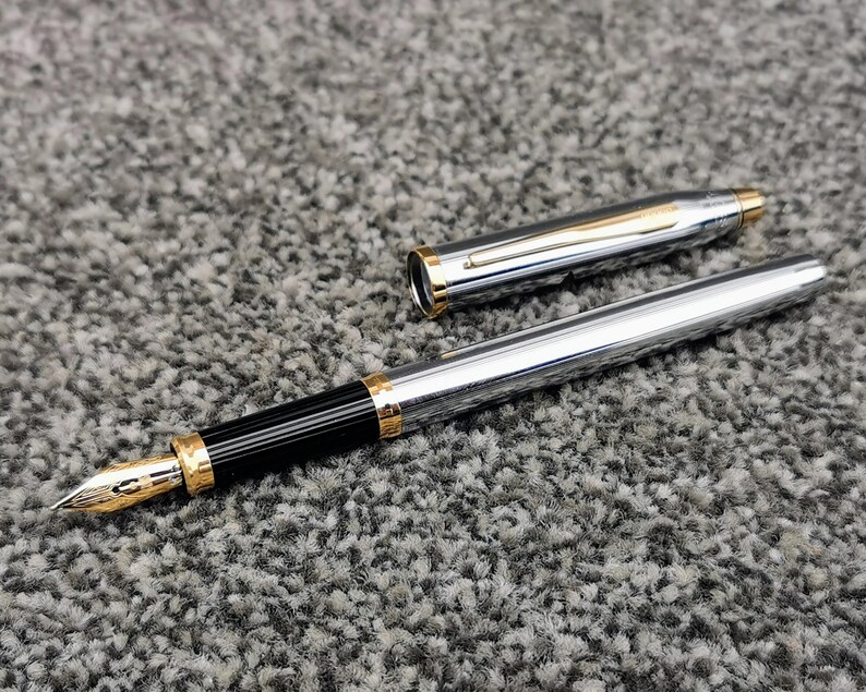 May include: A silver fountain pen with gold accents and a black grip. The pen is uncapped, revealing the gold nib. The cap is lying next to the pen. The pen is on a gray, textured surface.