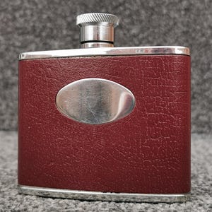 Può includere: Una fiaschetta rettangolare in metallo rivestita in similpelle testurizzata color rosso scuro. La fiaschetta presenta un tappo a vite in metallo argentato lucido e una placca metallica ovale e liscia centrata sulla parte anteriore.