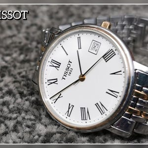 May include: A Tissot 1853 wristwatch with a white dial, black Roman numeral hour markers, and a date window at the three o'clock position. The watch features a two-tone silver and gold-colored metal link bracelet and a gold-colored crown.