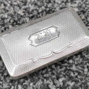 May include: A rectangular silver-toned metal case featuring a fine engine-turned wavy line pattern across the surface. The centre displays a smooth, decorative cartouche with the engraved initials "ED" in a stylised, ornate script font.