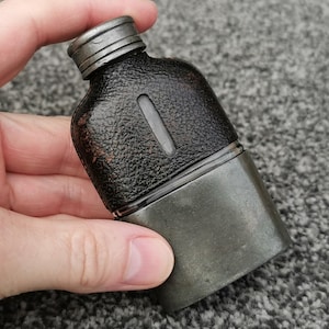 以下が含まれることがあります： 黒い革の質感のあるボディと金属製のベースとキャップが付いた、小さなアンティークフラスコ。 フラスコは長方形で、小さな透明な窓があります。 金属部品は暗く、古びた外観をしています。