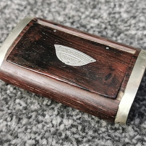 May include: A rectangular wooden box with a dark brown finish and silver metal trim. The box features a small, silver emblem with the text "LA SOURCE DEPOSE" on the top. The wood grain is visible.
