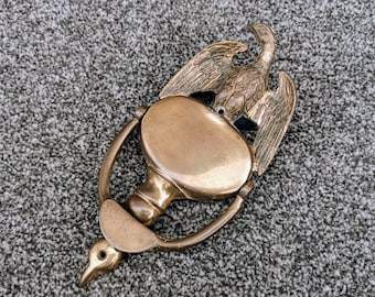 May include: A brass door knocker shaped like an eagle with its wings spread. The eagle is perched on top of a round, brass knocker with a hole in the center. The knocker is attached to a brass ring.