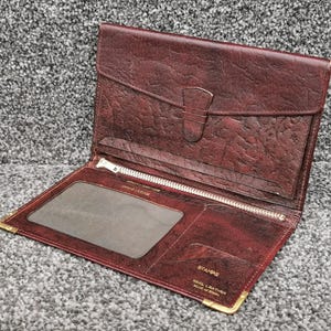 May include: Open burgundy leather wallet with a flap closure and a silver zip. The wallet has a clear ID window and a small pocket. The corners are reinforced with gold-coloured metal.