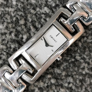Op de afbeelding: Een zilverkleurig rechthoekig polshorloge met een witte wijzerplaat met een subtiel textuurpatroon en het DKNY-logo. Het horloge heeft een gepolijste metalen schakelarmband met een geometrisch ontwerp en eenvoudige zwarte wijzers.