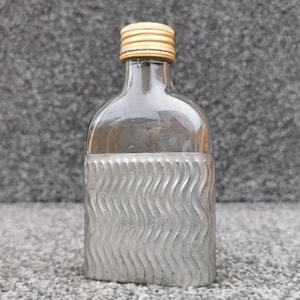 Pode incluir: Uma garrafa de vidro transparente com ombros arredondados e gargalo estreito, rematada por uma tampa de rosca dourada e estriada. A metade inferior é revestida por uma manga de metal prateado com um padrão de textura ondulada vertical.