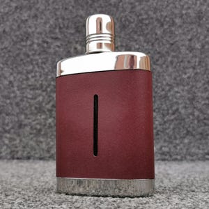 Könnte beinhalten: Ein rechteckiger Flachmann aus Edelstahl mit einer dunkel-weinroten, lederartigen Ummantelung. Der Flachmann hat einen polierten, silberfarbenen Metallverschluss mit Schraubkappe und ein vertikales Sichtfenster mittig auf der Vorderseite.