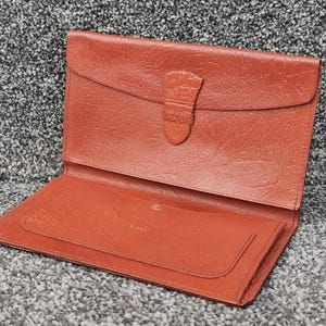 May include: Open, rust-colored leather wallet with a flap closure and a small pocket. The wallet has a textured surface and a buckle closure. The interior features a smaller pocket and a gold-colored logo.
