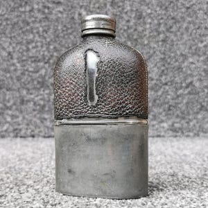 Könnte beinhalten: Ein Metallflachmann mit einem dunkelbraunen, strukturierten Lederüberzug im oberen Bereich, der eine vertikale Aussparung aufweist, die das silberfarbene Metall darunter freilegt. Der untere Teil besteht aus glattem, mattem Silbermetall.