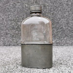 Peut inclure: Une flasque en verre transparent avec un haut arrondi et un bouchon métallique à vis, dotée d'une base en métal gris foncé qui recouvre la partie inférieure du récipient. La flasque présente un profil plat et ovale au design utilitaire.