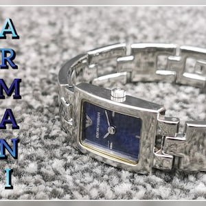 May include: A silver-toned Armani watch with a rectangular blue face and a matching link bracelet. The watch features the Armani logo and is set against a textured gray background. The word "Armani" is displayed vertically in blue letters.