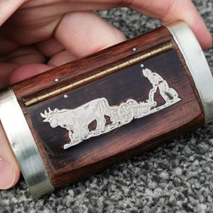 Pode incluir: Uma caixa retangular de madeira com bordas de metal prateado e um relevo decorativo prateado. O relevo mostra um boi puxando uma carroça com uma pessoa empurrando. A madeira é marrom escuro com grão visível.