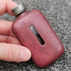 Könnte beinhalten: Ein rechteckiger Taschenflachmann, der mit einem strukturierten, dunkelweinroten, lederartigen Material überzogen ist. Der Flachmann verfügt über ein schmales, vertikales Sichtfenster aus Glas in der Mitte und einen metallischen Schraubverschluss.