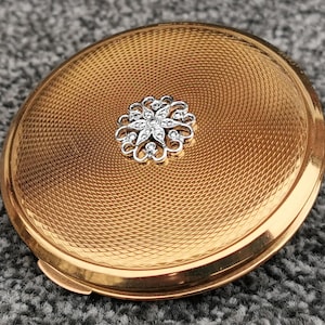 May include: Oval gold-toned compact with a textured surface and a silver-colored floral embellishment. The compact has a hinged opening and a polished gold rim. The design features a spiral pattern radiating from the center.