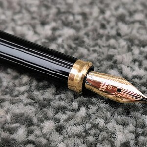 May include: Close-up of a black and gold fountain pen. The pen has a black barrel with gold accents and a gold nib. The nib is engraved with the word "BOSS" and decorative patterns. The pen is on a gray surface.