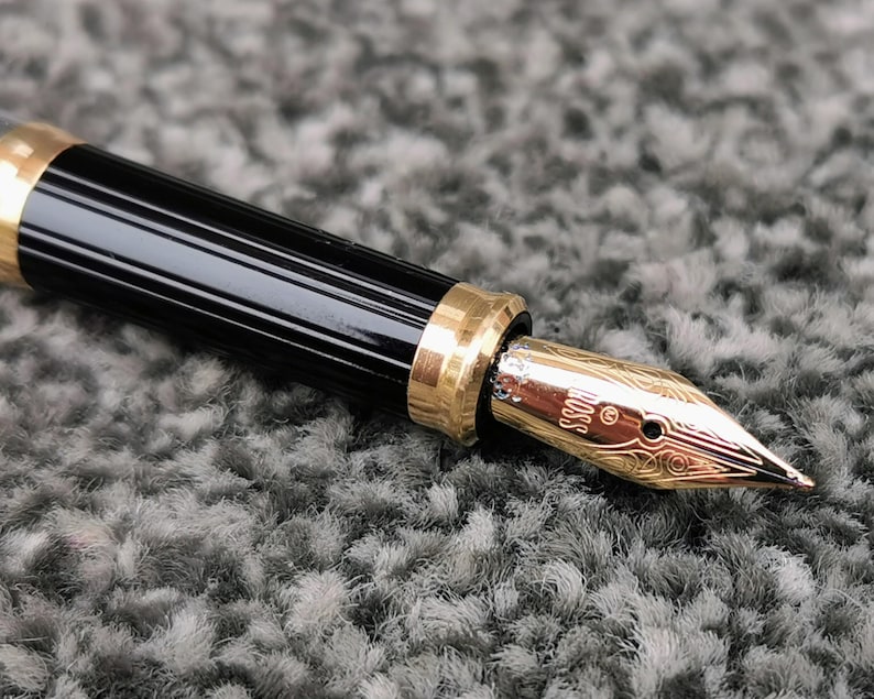May include: Close-up of a black and gold fountain pen. The pen has a black barrel with gold accents and a gold nib. The nib is engraved with the word "BOSS" and decorative patterns. The pen is on a gray surface.