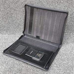 May include: Open black leather portfolio with a zippered closure. The interior features a document holder, card slots, and a small notepad. The portfolio is designed for professional use and has a textured finish.