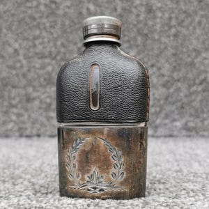 Könnte beinhalten: Ein Taschenflachmann mit einem silberfarbenen Metallsockel, der ein eingraviertes Lorbeerkranz-Motiv aufweist, und einem mit schwarzem Leder umwickelten Oberteil. Der Flachmann besitzt ein kleines vertikales Sichtfenster und einen Schraubverschluss.