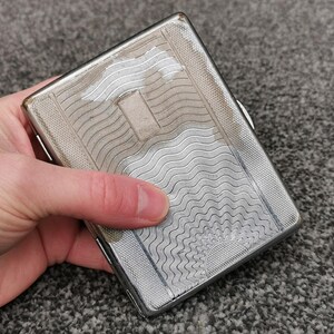 Könnte beinhalten: Eine silberne Zigarettenetui mit einem strukturierten, wellenartigen Muster. Das rechteckige Etui hat einen Klappdeckel und einen kleinen rechteckigen Akzent. Das Etui wird in einer Hand gehalten und zeigt das komplizierte Design und den metallischen Glanz.
