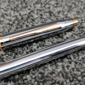 May include: A silver pen with gold accents, including a gold clip and trim. The pen's body has a textured, ridged design. The pen is partially disassembled, with the cap and body separated, revealing the pen's inner components. The pen is on a gray, textured surface.