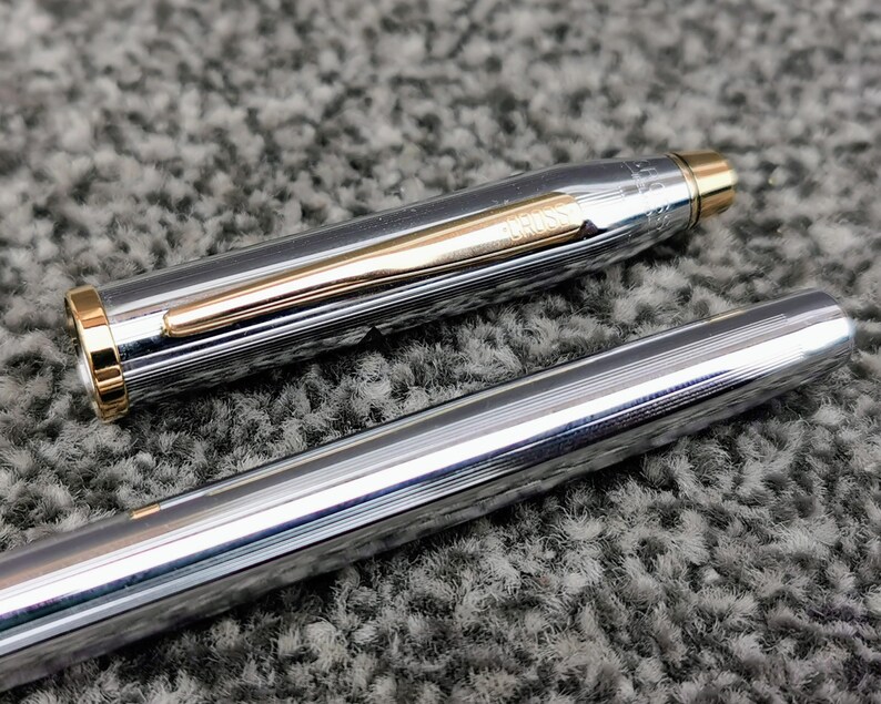 May include: A silver pen with gold accents, including a gold clip and trim. The pen's body has a textured, ridged design. The pen is partially disassembled, with the cap and body separated, revealing the pen's inner components. The pen is on a gray, textured surface.
