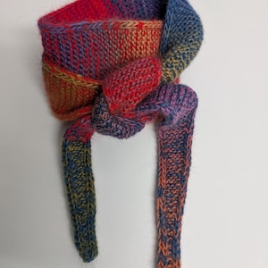 Può includere: Una sciarpa lavorata a mano a maglia in una vibrante gamma di colori, tra cui rosso, blu, arancione e giallo. La sciarpa è annodata al centro e presenta un design intrecciato strutturato. La sciarpa è realizzata in filato morbido.