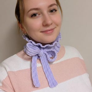 Op de afbeelding: Een lila gebreide halsdoek met een strik. De sjaal heeft een gerimpelde kraag en lange banden. De persoon op de foto draagt een roze en wit gestreepte trui.