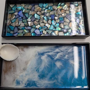 May include: Two rectangular black trays. The top tray features a mosaic of iridescent blue, white, and silver shell fragments embedded in resin. The bottom tray displays a swirling blue and white resin pattern with a single white seashell attached.