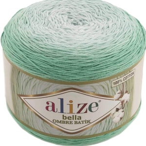 May include: A ball of Alize Bella Ombre Batik yarn, transitioning from light blue to green. The label states "100% Cotton" and features the Alize logo and product name. Ideal for knitting and crochet projects.