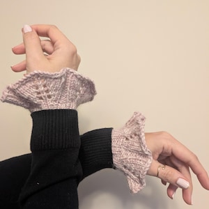 May include: A pair of light pink knitted wrist cuffs featuring a delicate ruffled edge and an openwork lace pattern. The cuffs are worn over the sleeves of a black long-sleeved garment, showcasing a textured knit construction and a flared design.