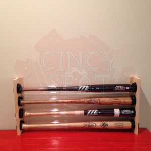 May include: A wooden baseball bat display rack with four clear tubes holding baseball bats. The bats are signed by Austin Kearns,  Lou Brock,  Devin Mesoraco, and  Johnny Bench. The rack is on a red surface.