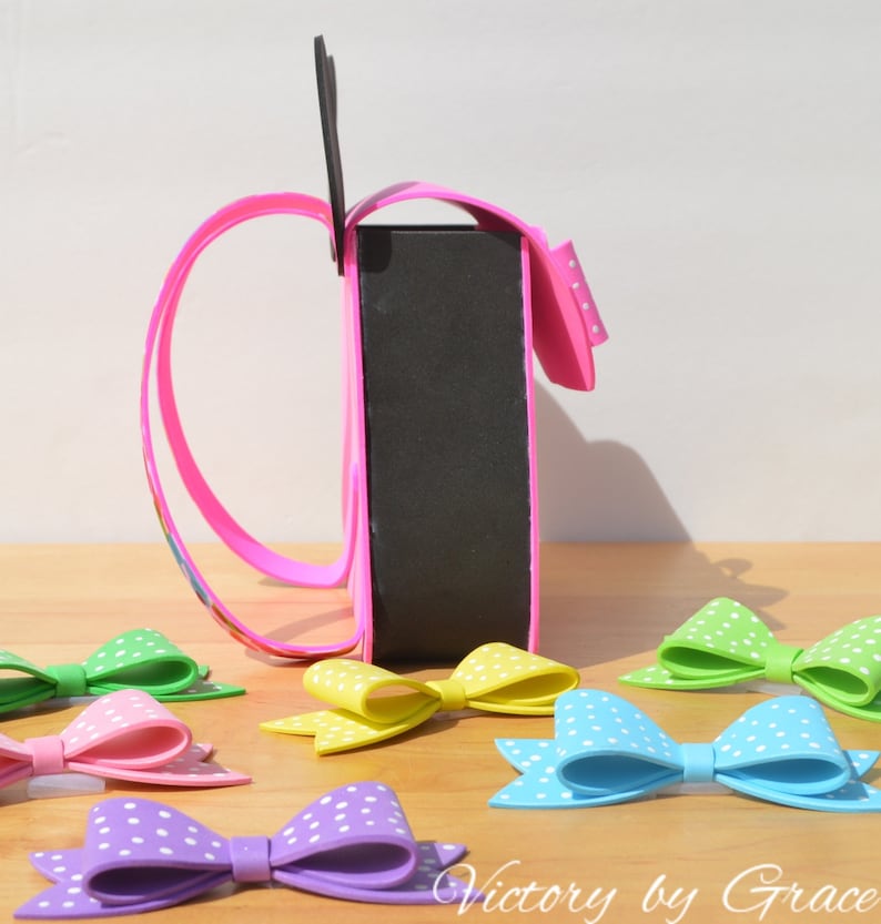 Puede incluir: Una bolsa de artesan&iacute;a de espuma rosa con un asa de espuma negra y lunares blancos. La bolsa est&aacute; sentada sobre una superficie de madera con varios lazos de espuma de colores con lunares blancos.