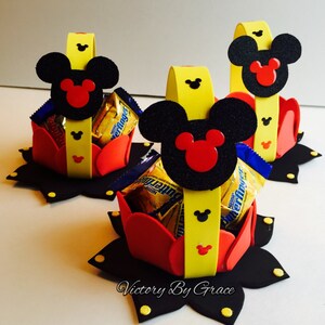 May include: Three Mickey Mouse themed party favor baskets made of red, yellow, and black foam. Each basket has a Mickey Mouse head with black ears and red inner ears. The baskets are filled with small chocolate candy bars.