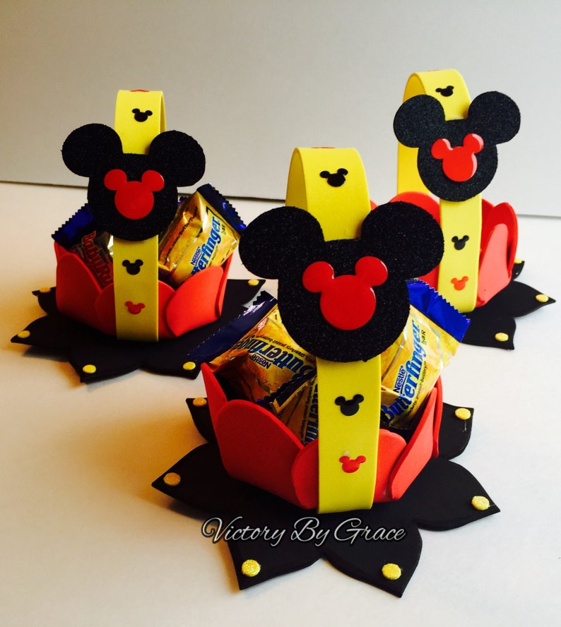 May include: Three Mickey Mouse themed party favor baskets made of red, yellow, and black foam. Each basket has a Mickey Mouse head with black ears and red inner ears. The baskets are filled with small chocolate candy bars.