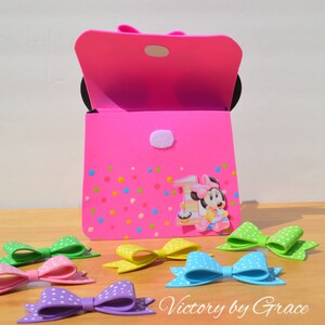 Puede incluir: Una bolsa de fiesta de espuma rosa con un personaje de Minnie Mouse y el n&uacute;mero uno en ella. La bolsa est&aacute; decorada con lunares de colores. Hay varios lazos de lunares de colores sobre una superficie de madera delante de la bolsa.