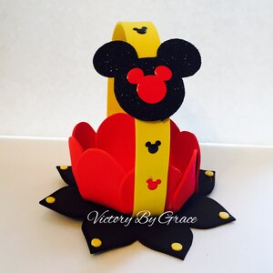 May include: A red and yellow Mickey Mouse themed party favor basket. The basket is made of foam and has a yellow handle with a black Mickey Mouse head on top. The basket is decorated with black and yellow polka dots.