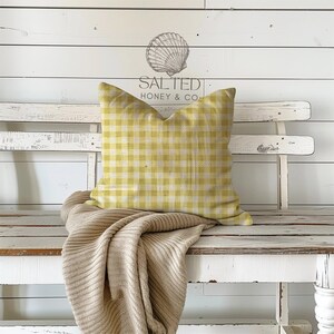 May include: A yellow and white gingham patterned pillow rests on a white bench, with a beige knitted throw draped over it. The background is a white wooden wall with the text "SALTED HONEY & CO" and a seashell illustration.