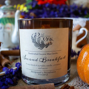 May include: A brown glass candle jar with a white label that reads "Handcrafted Coconut Wax Candle Second Breakfast A cozy blend of pumpkin, vanilla, cinnamon and pecan Handpoured in Pennsylvania 8 oz (226 g)"