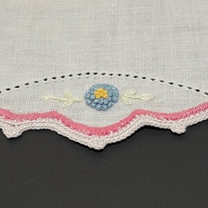 May include: A white linen handkerchief with a pink and white scalloped edge and a blue and yellow embroidered flower.