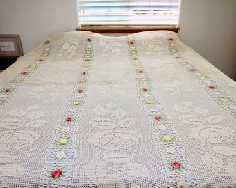 May include: A white crocheted bedspread with a floral design. The bedspread has a delicate pattern of roses and leaves, with accents of pink, yellow, and green.