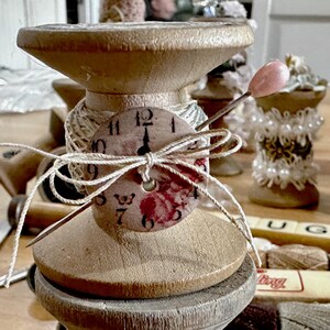 May include: A vintage wooden spool with a clock face attached, adorned with twine and a needle with a pink head. The spool is part of a collection of decorative items, including other spools and embellished objects, suggesting a craft or vintage theme.