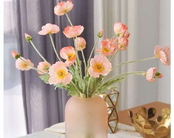 May include: A bouquet of pink silk poppy flowers in a frosted pink glass vase. The vase is on a white tablecloth.