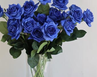 May include: A bouquet of vibrant blue roses in a clear glass vase. The roses have multiple petals and green leaves. The vase is filled with water and the stems of the roses are visible. The background is a plain white wall.